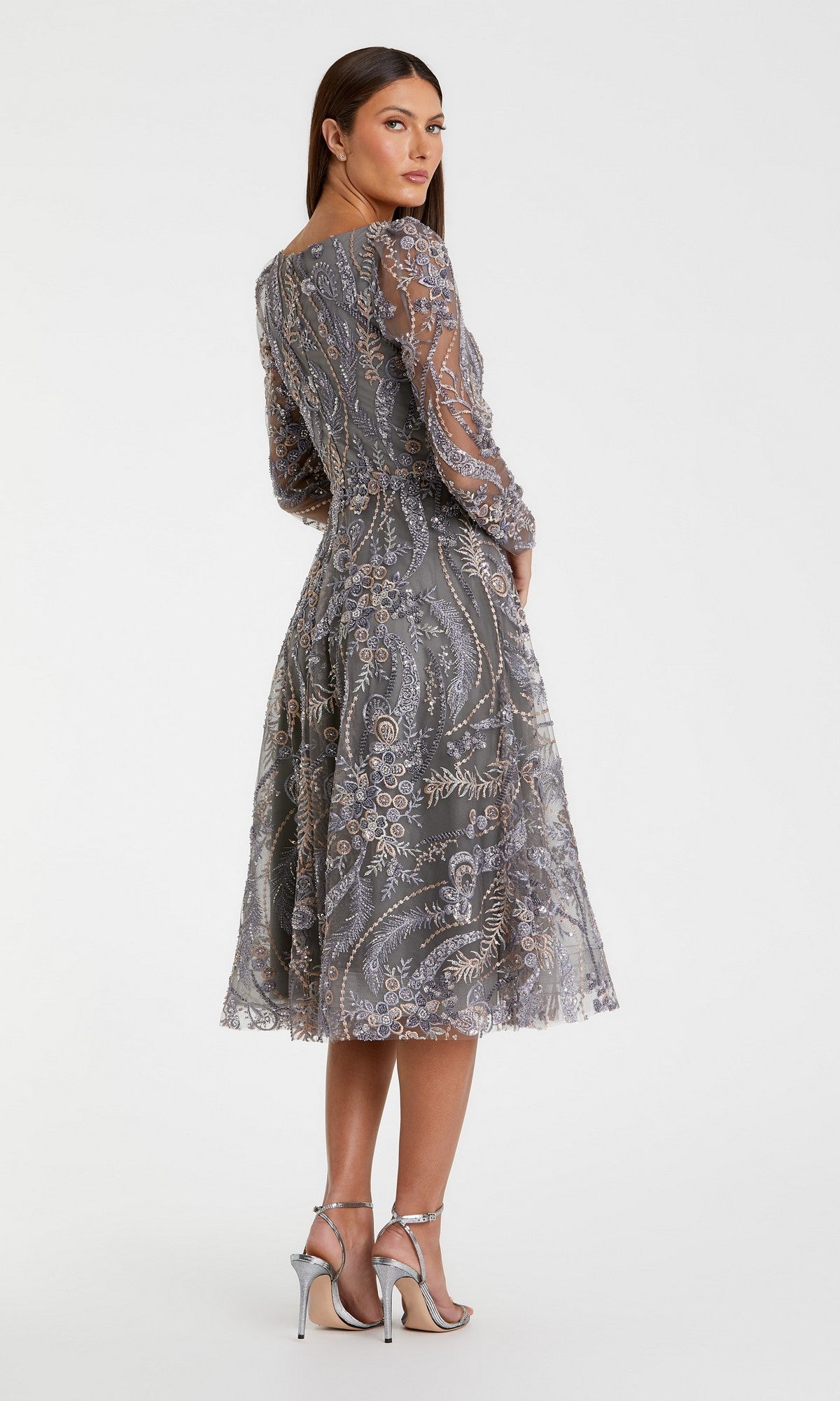 A woman wearing the Mac Duggal 68913 Midi Party Dress—a silver, sequin-beaded dress featuring embroidered long sleeves and sheer accents—stands in high heels, slightly turned away against a plain white background.